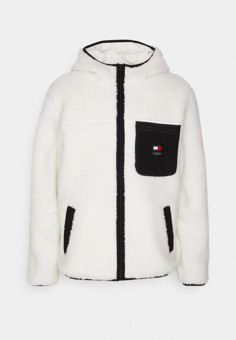 Veste en polaire blanche avec fermeture éclair, capuche et accents noirs. Comprend une poche frontale zippée et deux poches latérales. Texture douce.