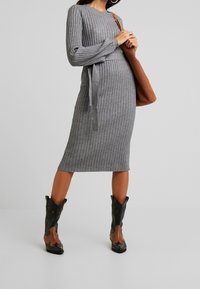Abito grigio a coste in maglia con maniche lunghe e cintura da annodare. Abbinato a stivali da cowboy neri e marroni con un design dettagliato. Borsa a spalla marrone.