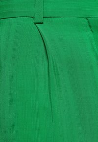 Gros plan sur un pantalon en tissu vert vif montrant la ceinture, les passants de ceinture et les plis avant.