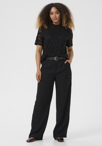 Top corto a maniche corte in pizzo nero con collo alto, abbinato a pantaloni neri a gamba larga e una cintura nera. L'outfit comprende scarpe nere lucide.