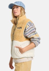 Vesti tan dan krim dari sherpa dengan resleting di depan, dilengkapi logo dan saku samping. Dipakai di atas lengan panjang bermotif dan topi denim.