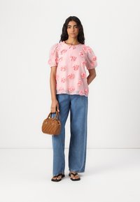 Blusa rosa chiaro con applicazioni a forma di rosa, maniche corte a sbuffo; abbinata a pantaloni in denim blu larghi e una piccola borsa intrecciata marrone.