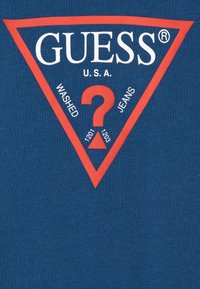 Tmavě modré tričko s velkým červeným trojúhelníkovým logem a bílým textem "GUESS U.S.A." a otazníkem, doplněné nápisem "WASHED JEANS" a čísly stylu.
