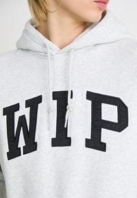 Grå hettegenser med snor, med store svarte "WIP" -bokstaver. Tøyer ser myke ut, med kontrasterende sømmer rundt bokstavene.