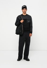 Hanorac negru cu un buzunar pe piept, asortat cu pantaloni cargo negri. Modelul poartă o geantă texturată neagră și o șapcă neagră.