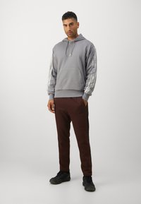 Szürke kapucnis pulóver elülső zsebbel és húzózsinórral, az ujjakon ezüst csíkokkal. Barna nadrág fekete részletekkel és logóval. Fekete cipők.