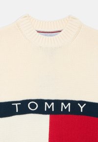 Suéter de punto color crema con cuello redondo acanalado y una banda azul marino con la palabra "TOMMY". Un bloque rojo añade un contraste de color.