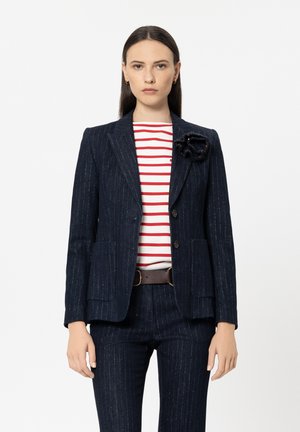 Marineblaues Nadelstreifen-Sakko mit aufgesetzten Taschen, kombiniert mit einem rot-weiß gestreiften Hemd. Akzentuiert durch eine schwarze Stoffblume am Revers.