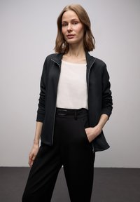 Giacca nera con cerniera e texture liscia, abbinata a una camicia beige chiaro e pantaloni neri su misura con dettagli di cintura.