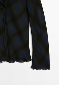 Chemise à manches longues à carreaux foncés avec des bords effilochés le long de l'ourlet, des poignets et de la patte de boutons sur fond blanc.
