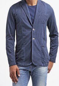 Uomo che indossa un blazer casual blu navy con due bottoni sopra una t-shirt blu navy e jeans azzurri, in piedi davanti a uno sfondo bianco.