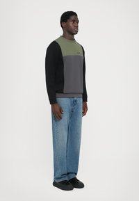 Sweatshirt color-blockat cu secțiuni de verde măsliniu, gri și negru. Prezintă un guler rotund și detalii cu logo. Asortat cu blugi largi albaștri.