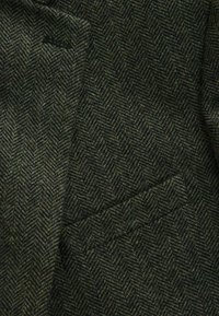 Hanfgewebe im Fischgrätmuster in dunklen Grüntönen und Schwarz, mit einer strukturierten Oberfläche, die eng gewobene Fasern und ein markantes Zickzackdesign zeigt.