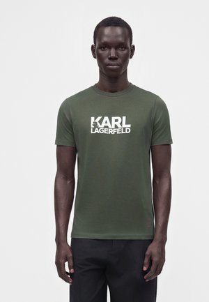Mand iført en olivengrøn kortærmet t-shirt med teksten "KARL LAGERFELD" i hvid, stående mod en ensfarvet hvid baggrund.