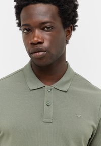 Zielona koszulka polo z teksturowanej tkaniny, z wąskim dekoltem na dwa guziki i małym haftowanym logo po lewej stronie klatki piersiowej. Krótkie rękawy.