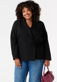 Zwarte blazer in wikkelstijl met lange mouwen en een striksluiting, gecombineerd met lichtblauwe jeans en een bordeauxrode handtas. Glad materiaal, zonder patronen.