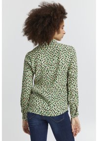 Blusa a maniche lunghe in tessuto leggero, caratterizzata da un motivo astratto verde e nero su sfondo beige, con un orlo leggermente arrotondato.