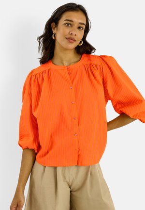 Mujer con blusa abotonada de rayas finas en color naranja brillante con mangas abullonadas y pantalones beige de talle alto, de pie con una mano en la cadera.