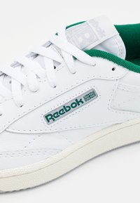 Zapatilla de cuero blanca con acentos verdes, que cuenta con una suela texturizada, cordones planos y el logo de Reebok en el lateral y la lengüeta.