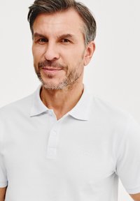 Biała koszulka polo z kołnierzykiem, dwoma guzikami oraz dyskretnym logo marki. Gładki materiał i klasyczny krój.