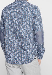Homme portant une chemise à manches longues bleu floral avec des coudières grises et un pantalon gris clair, se tenant avec une main dans la poche.