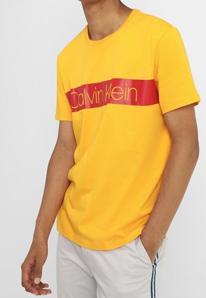 Osoba nosząca jaskrawożółtą koszulkę Calvin Klein z czerwoną poziomą paską z logo, w połączeniu z jasnymi spodniami z bocznymi paskami.