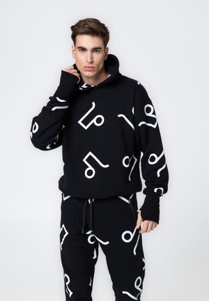Młody mężczyzna ubrany w czarną bluzę z kapturem i spodnie z białymi abstrakcyjnymi wzorami nut muzycznych, stojący na tle jednolitego białego tła.