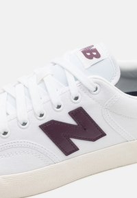 Fehér vászon sneaker lila bőr 'N' logóval, lapos fehér fűzőkkel és strukturált gumitalppal. Steppelt díszítések és megerősített fűzőlyukak jellemzik.