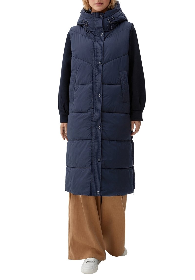 Femme portant une longue doudoune sans manches bleu marine avec capuche, un haut noir à manches longues et un pantalon large marron avec des baskets blanches.