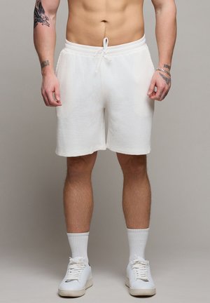 Männlicher Oberkörper und Beine, bekleidet mit weißen, strukturierten Shorts, weißen Socken, weißen Turnschuhen, mit Tattoos auf den Armen und einem Armband am rechten Handgelenk.