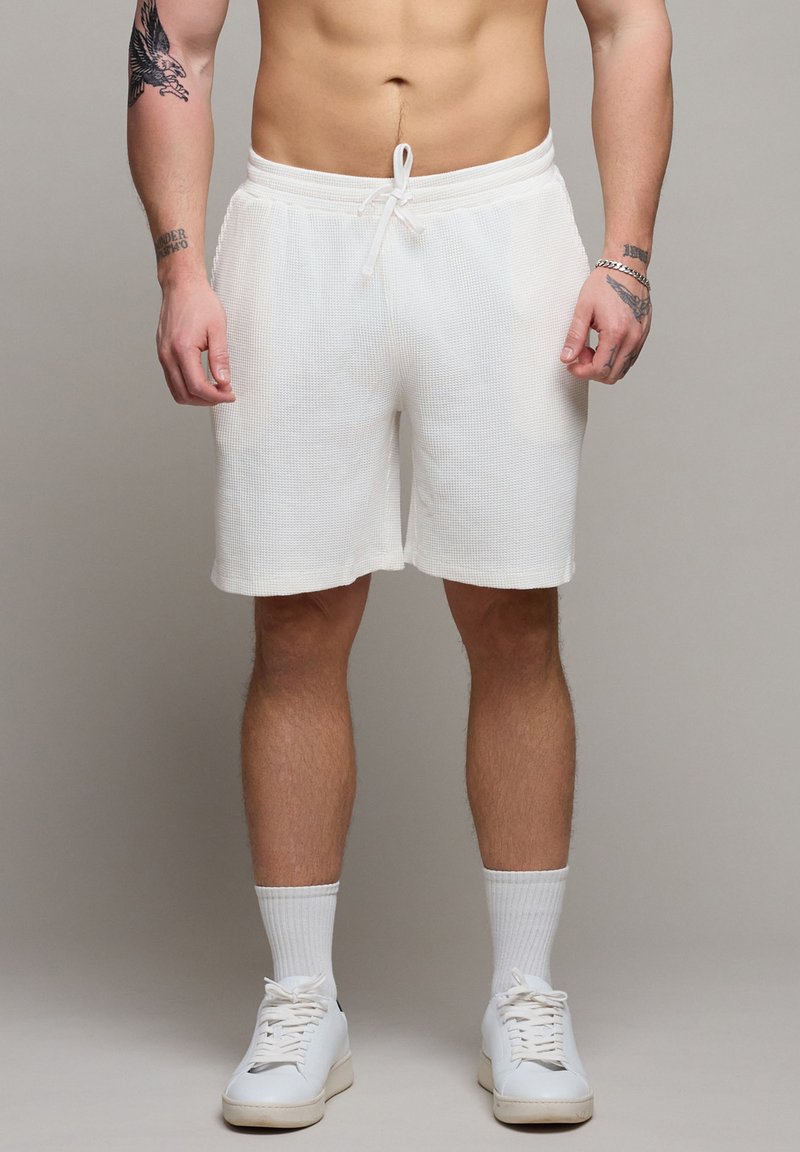 Torso et jambes d'un homme portant un short texturé blanc, des chaussettes blanches, des baskets blanches, avec des tatouages sur les bras et un bracelet au poignet droit.