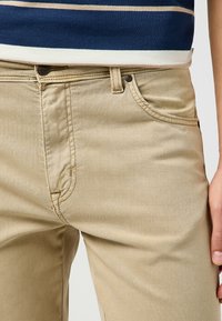 Chino in cotone beige con una superficie liscia, dotati di zip con bottone, tasche laterali e dettagli cuciti lungo le cuciture.