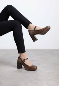 Scarpe con tacco alto in pelle marrone con punta tonda, cinturino alla caviglia e tacco spesso. Texture liscia con piattaforma a contrasto.