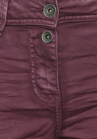 Gros plan sur un tissu bordeaux avec deux boutons en métal, dont un embossé avec "Cecil," et des coutures visibles sur la zone de la poche d'un vêtement.