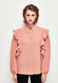 Blusa rosa con hombros con volantes y mangas abullonadas largas. Tejido ligero, con botones en la parte delantera, textura sutil, complementada con un cuello clásico.
