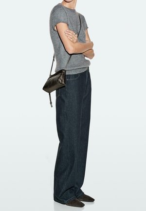 Person trägt graues Strickoberteil mit kurzen Ärmeln, weite dunkelblaue Jeans, schwarze Umhängetasche und dunkle spitze Schuhe, steht mit verschränkten Armen.