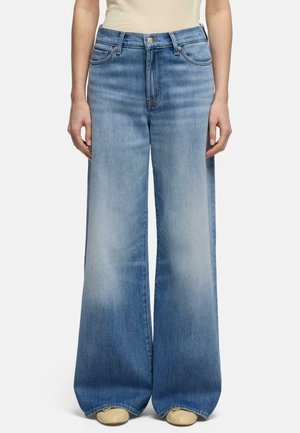 Person iført lyseblå jeans med vide ben, høj talje og falmede detaljer, kombineret med beige sko og en beige top.