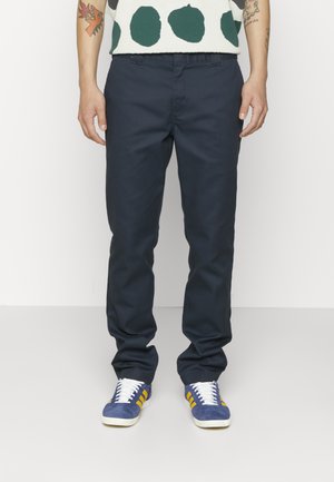 Hombre con pantalones azul marino, zapatillas azules con rayas amarillas y camisa con grandes lunares verdes, de pie con los brazos relajados.