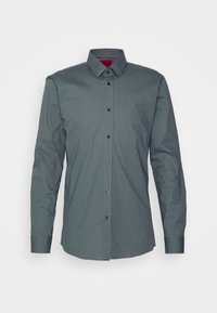 Camicia verde scuro a maniche lunghe in cotone. Presenta un colletto con bottoni, una patta anteriore e bottoni neri con un'etichetta rossa all'interno del colletto.