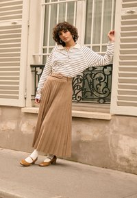 Camicia a maniche lunghe a righe con colletto, abbinata a una gonna beige plissettata con cintura. Scarpe marroni con tacco largo e calzini bianchi completano il look.