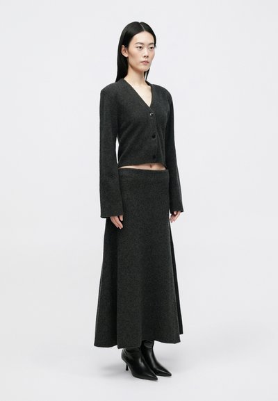 Γυναίκα που φοράει ένα σκούρο γκρι μακρύ μανί�κι γιακά κουμπωτό πλεκτό και αντίστοιχη φούστα μέχρι τον αστράγαλο με μαύρες γόβες με μυτερή μύτη, στέκεται.