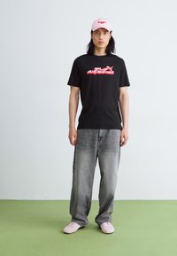Sort t-shirt med rød grafik, grå bredbenede jeans og lyserøde sneakers; iført en lyserød kasket. Ensfarvet baggrund med grøn gulv.