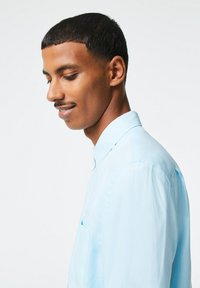 Camicia button-down azzurro chiaro in un tessuto morbido, con colletto, dettaglio della tasca e una vestibilità comoda, mostrata su un modello di profilo.