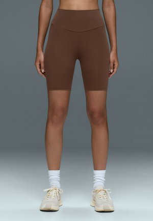 Kvinde iført højtaljede brune biker-shorts, hvide crew-sokker og beige sneakers, der står mod en ensfarvet grå baggrund.