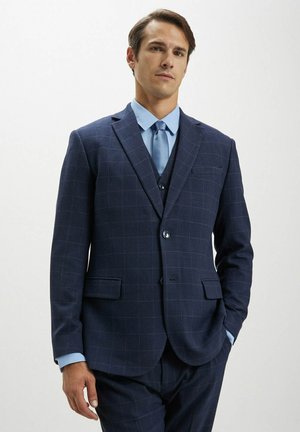 Jacket da suit a quadri blu navy con rever a pantaloni, chiusura a un bottone e due tasche frontali, abbinato a una camicia azzurra e una cravatta.