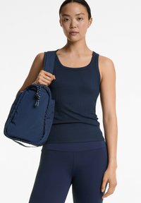 Marinefarbener, gerippter Tanktop mit rundem Ausschnitt, kombiniert mit dunkelblauen Leggings. Das Model hält einen gesteppten Rucksack mit Kordelzug.