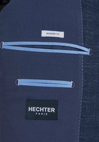 Marineblaue Stoff mit glatter Textur zeigt ein Etikett "MODERN FIT", eine Tasche mit hellblauen Reißverschlüssen und einem schwarzen Patch "HECHTER PARIS."