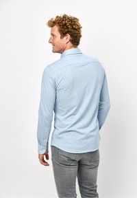 Camicia azzurra a maniche lunghe con colletto a punta, polsini abbottonati e una texture liscia, abbinata a jeans grigi, mostrata da dietro.