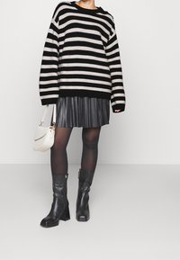 Schwarz-weiß gestreifter Pullover, schwarzer Faltenrock, blickdichte Strumpfhosen und schwarze Stiefel mit Absatz. Eine weiße Handtasche rundet das Outfit ab.