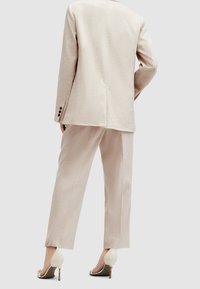 Beżowy lniany garnitur z dopasowaną marynarką i prostymi spodniami. Marynarka ma kieszenie z patką i subtelne detale w wentylacji z tyłu.